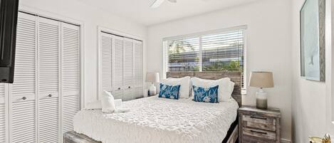 Un dormitorio con cama, mesitas de noche, ventilador de techo y ventana con persianas.