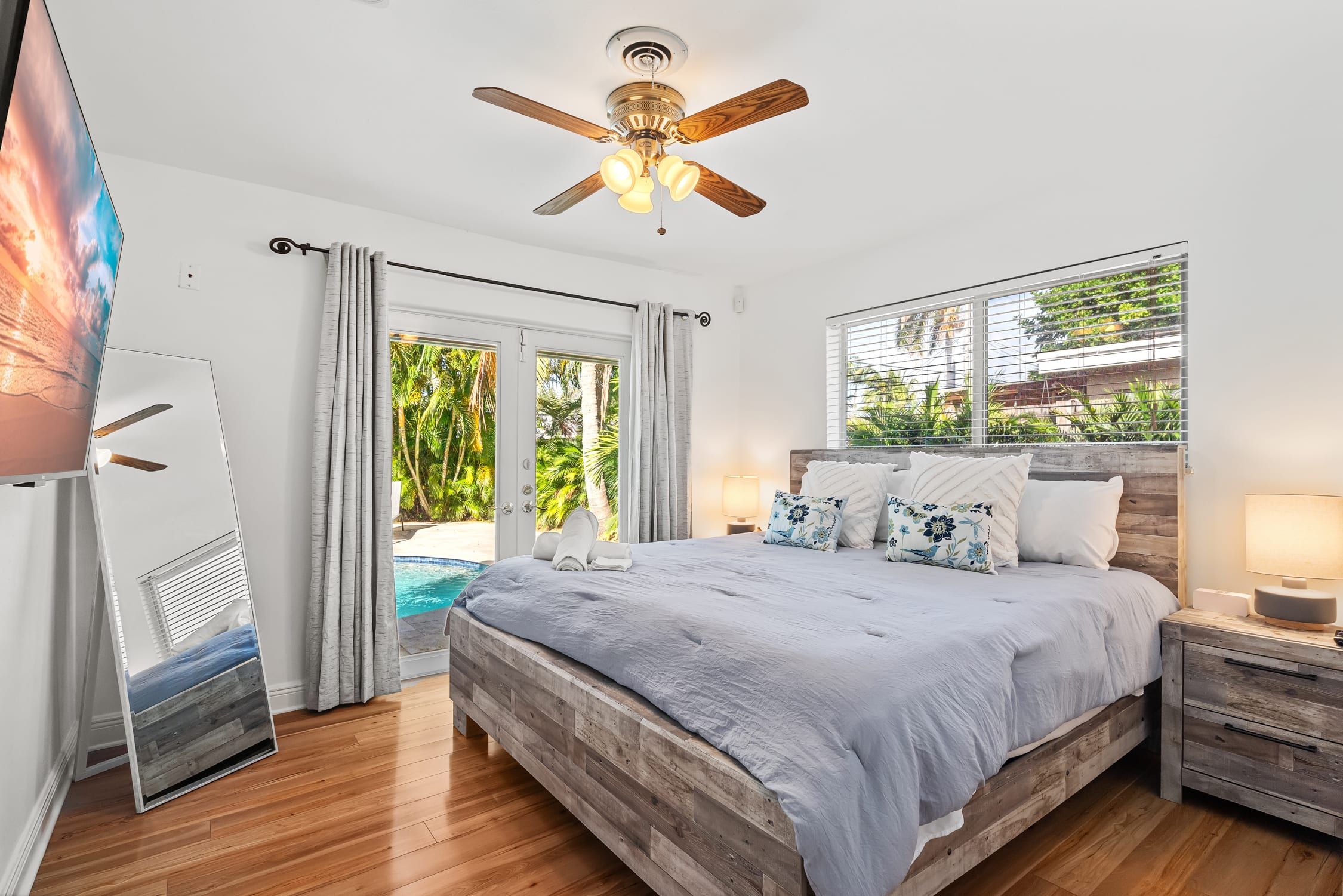 Une chambre avec un lit en bois, un ventilateur de plafond, une fenêtre avec des stores et une vue sur une piscine.