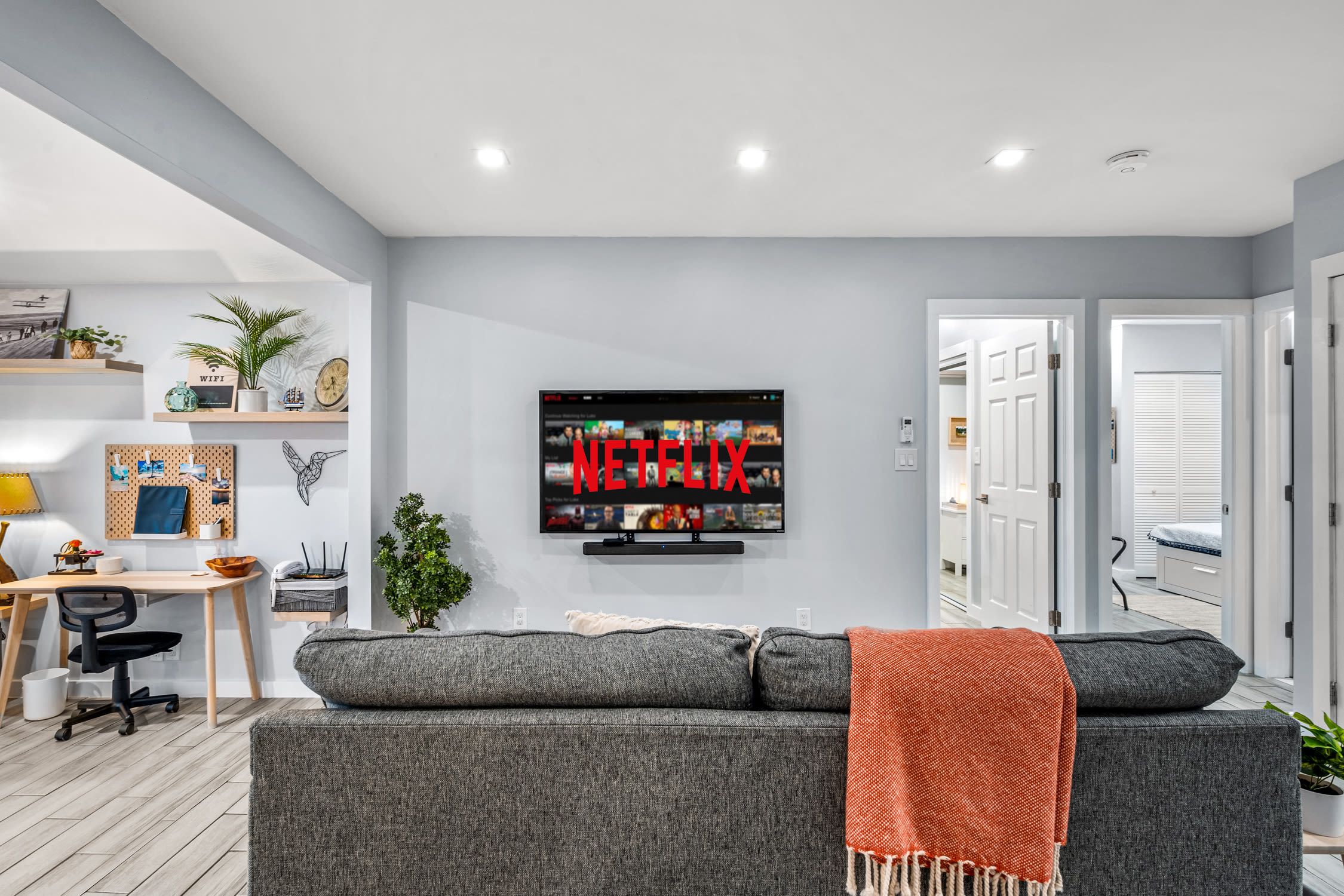 Een moderne woonkamer met een grijze bank, een flatscreen televisie aan de muur en een bureau met een computer.