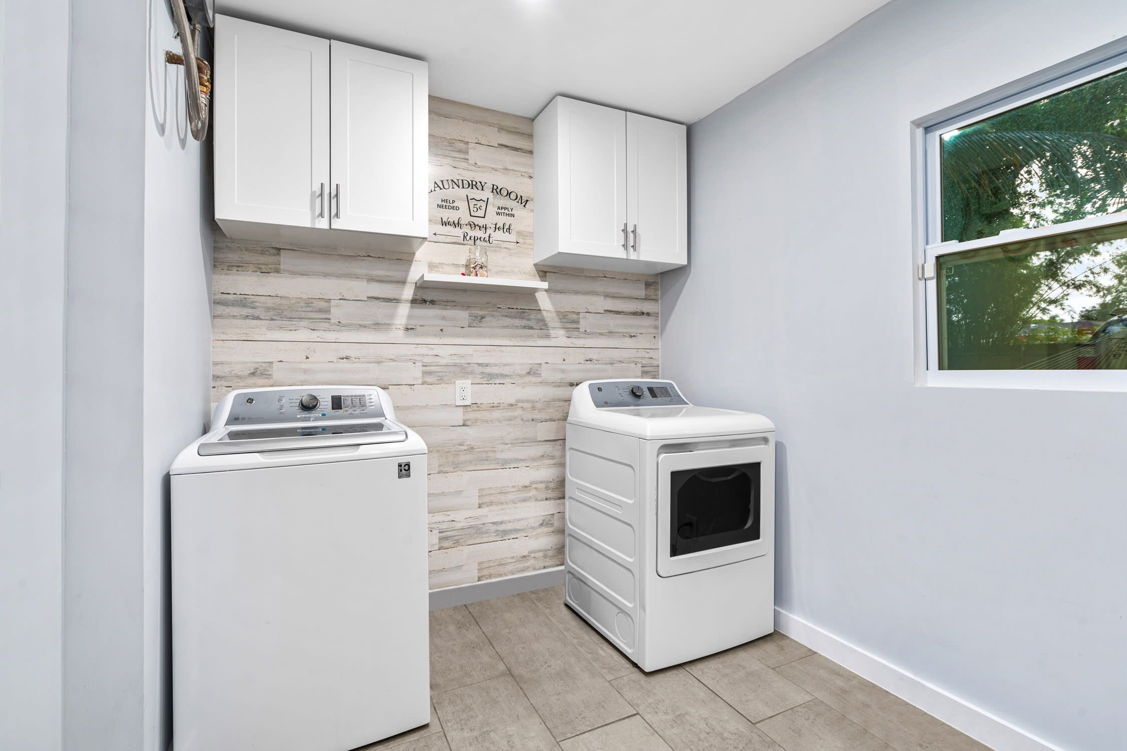 A laundry room with two white washing machines, a wooden wall with a laundry instruction sign, and a window with a view of trees.