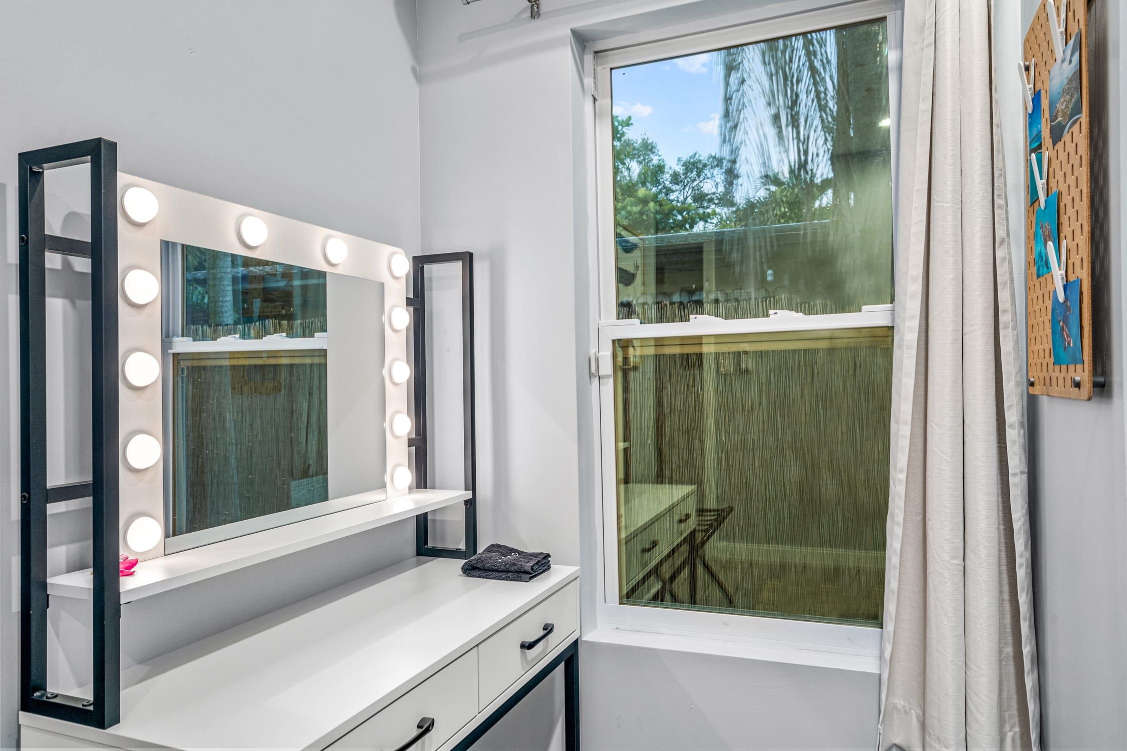 A room with a vanity mirror with lights, a window, and a wall-mounted pegboard with cloths.