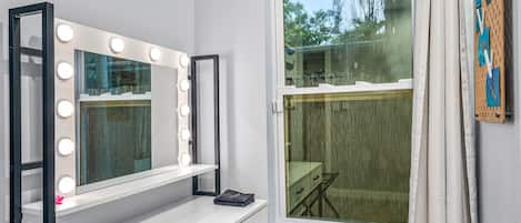 A room with a vanity mirror with lights, a window, and a wall-mounted pegboard with cloths.