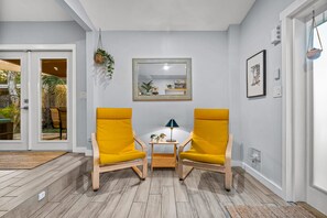 Een kamer met twee gele stoelen, een klein houten tafeltje, een spiegel en een deur naar een buitenruimte.