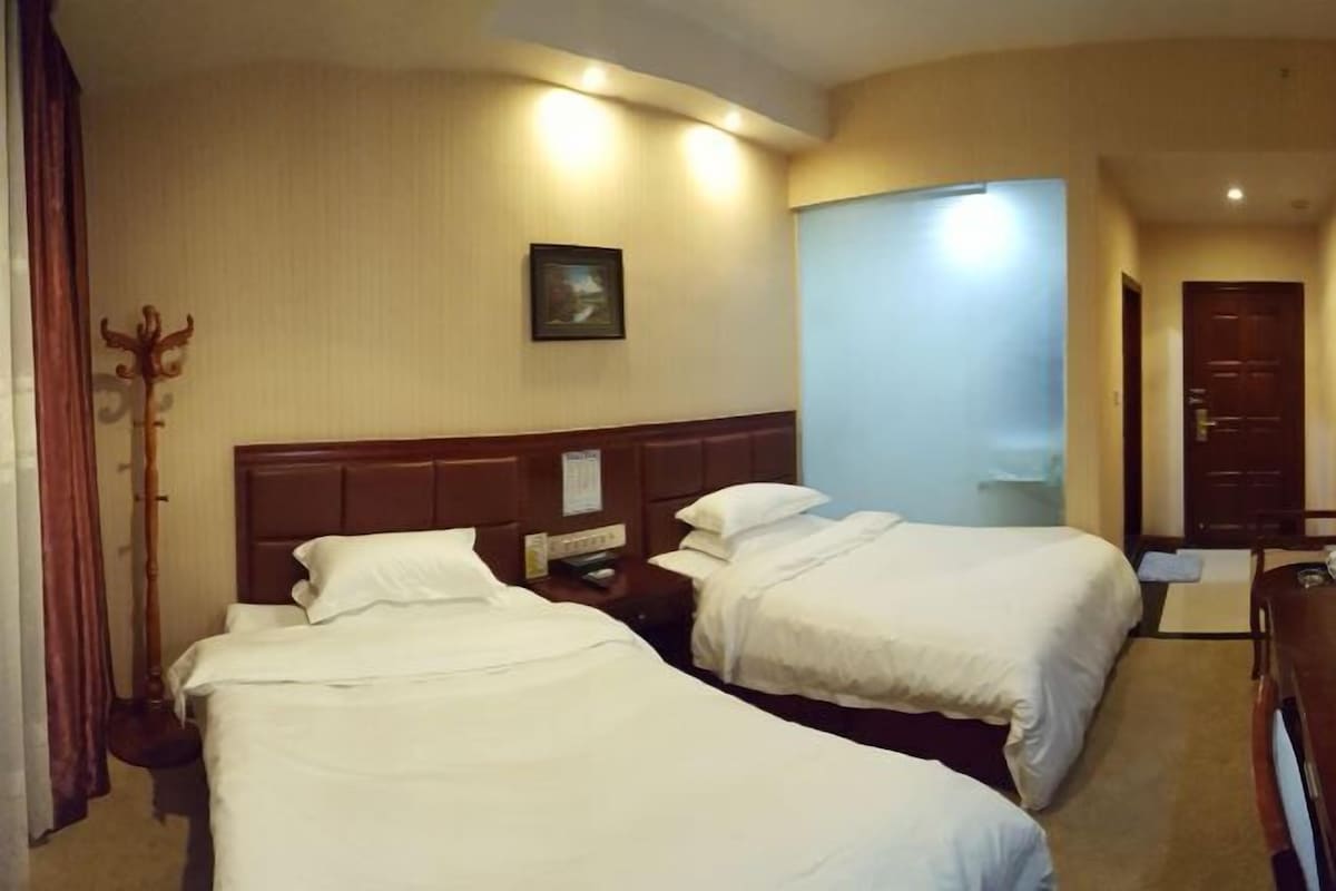 Una habitación de hotel con dos camas individuales, un cabecero de madera, una mesita de noche con teléfono, un perchero y una puerta que da a otra habitación.