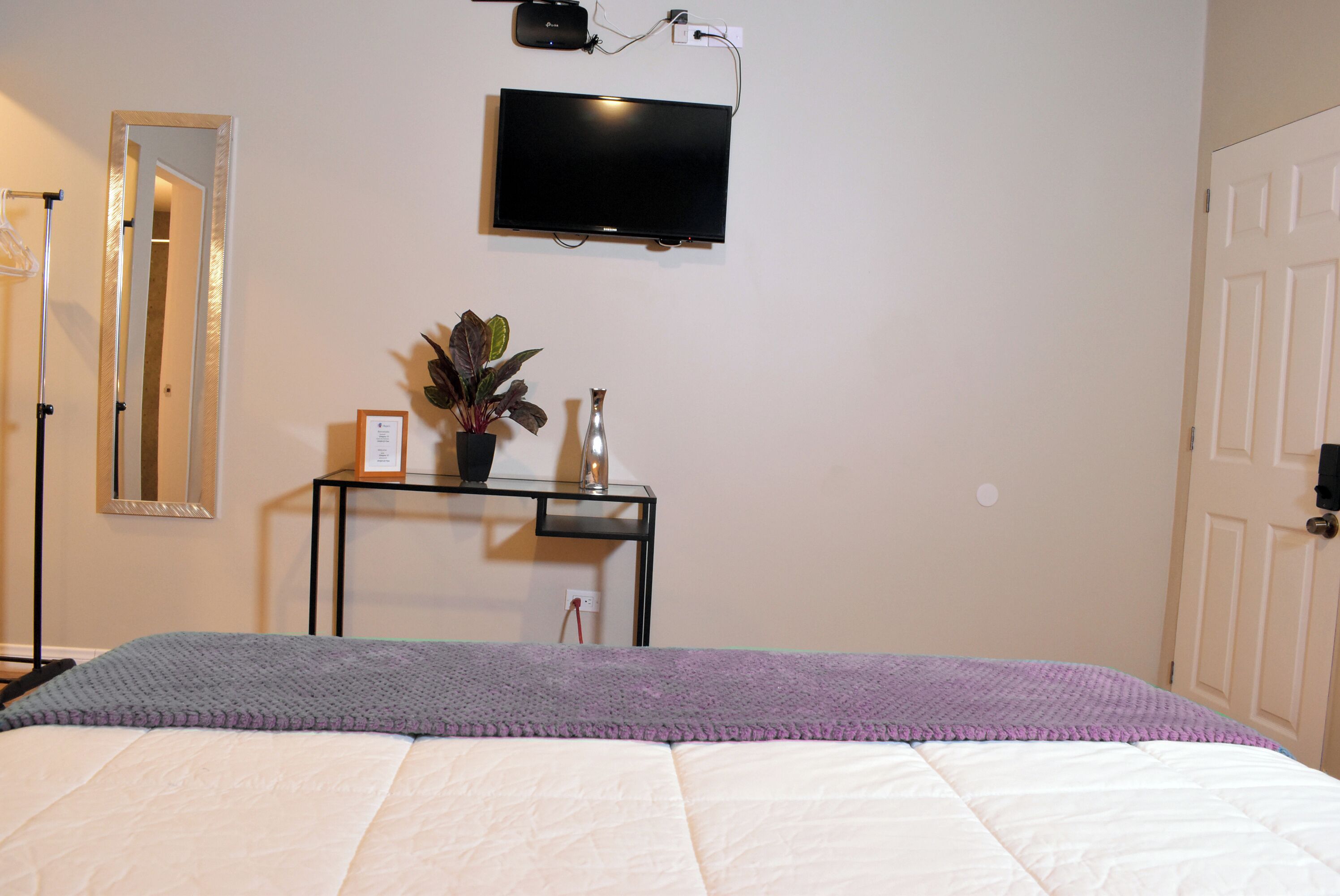 A bedroom with a bed, a TV mounted on the wall, a bedside table with a plant and a vase, and a mirror on the wall.