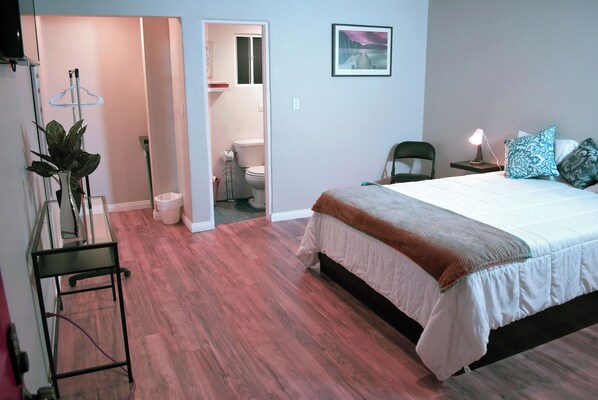 A bedroom with a bed, a nightstand, a chair, a plant, and a bathroom visible through an open door.
