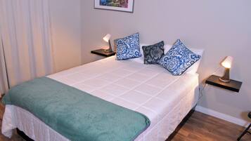 A neatly made bed with a green throw blanket, white bedding, and patterned pillows. There are two bedside tables with lamps, a framed picture on the wall, and a wooden floor.