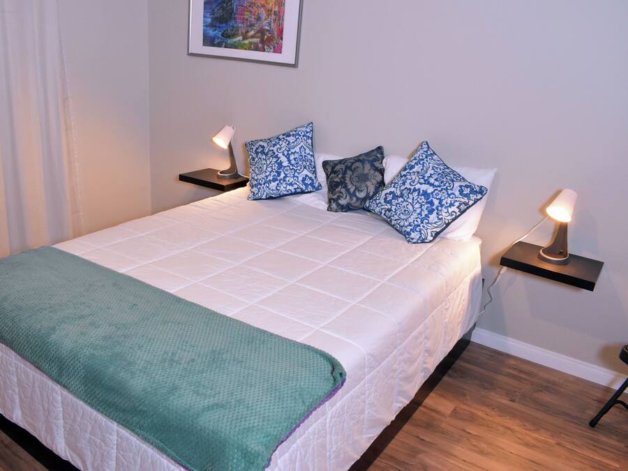 A neatly made bed with a green throw blanket, white bedding, and patterned pillows. There are two bedside tables with lamps, a framed picture on the wall, and a wooden floor.