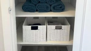A white shelving unit with three tiers; the top and middle tiers hold rolled towels in shades of gray and blue, while the bottom tier contains white baskets. - Beautiful house//self check in//7 mins to YEG Airport//Nature walk🌴 (Leduc)