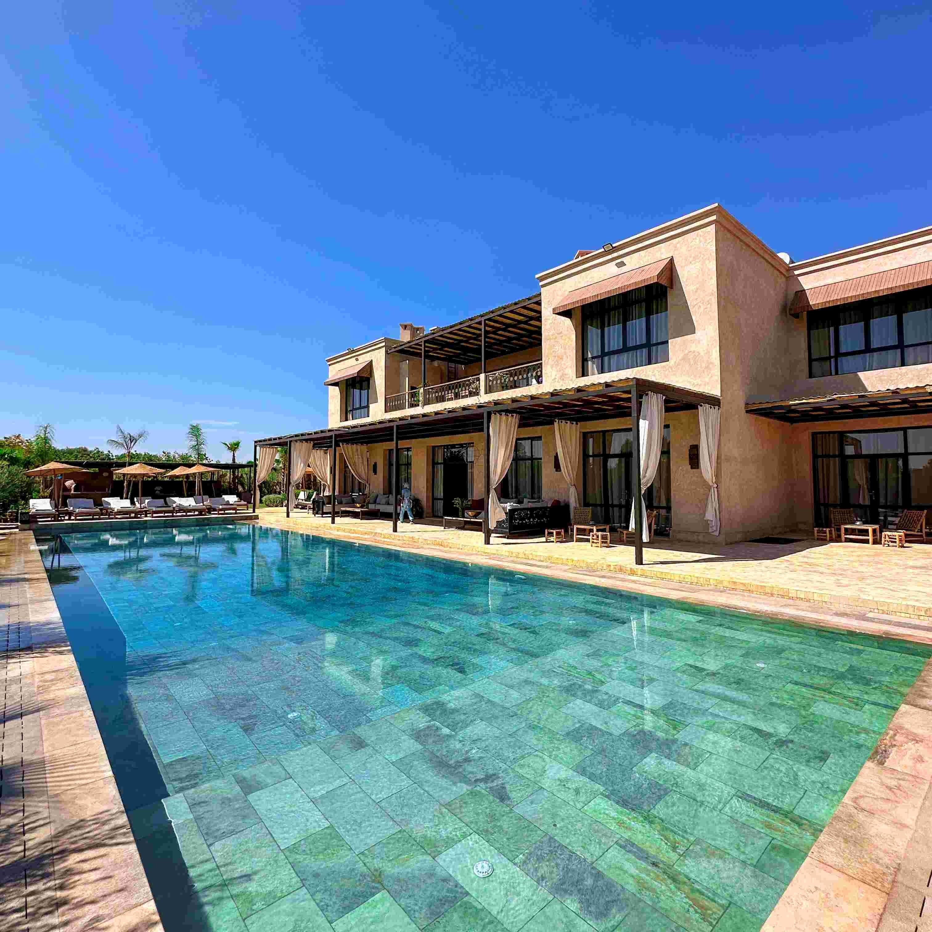A poolside area with a clear pool, lounge chairs, and a building with large windows and a covered patio.