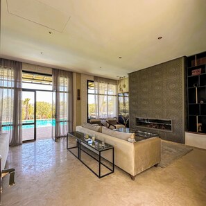 A spacious living room with a large sofa, a glass coffee table, and a fireplace. There is a view of a pool through large windows. - Al Destino Luxury Villa & Spa Marrakech (Oulad Hassoune)