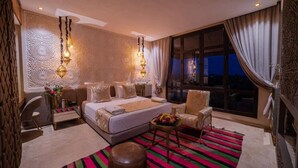 A modern hotel room with a large bed, a sitting area with a chair and a small table, a desk, and a large window offering a view of the night sky. - Al Destino Luxury Villa & Spa Marrakech (Oulad Hassoune)
