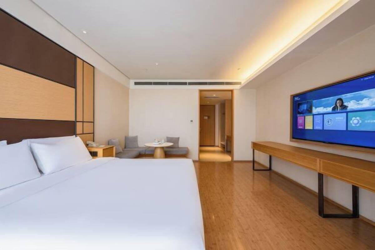 A modern hotel room with a large bed, a TV mounted on the wall, a small seating area, and a wooden bench.