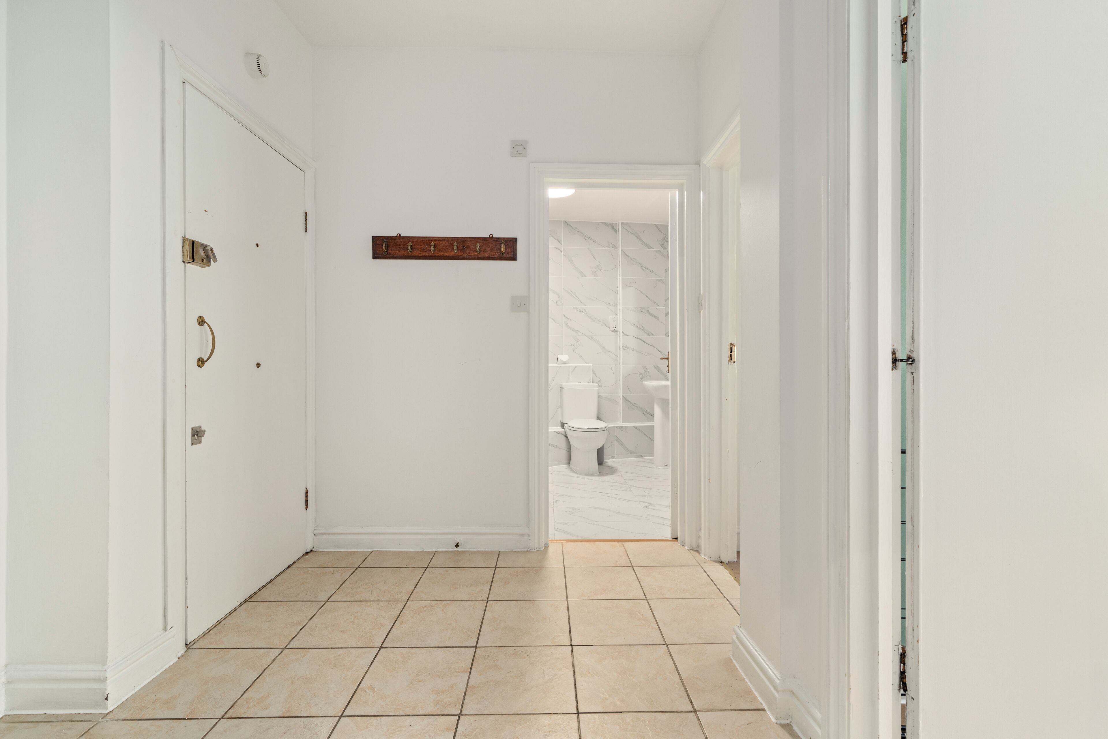 A hallway with white walls, a tiled floor, and a door leading to a bathroom with a toilet and marble flooring.