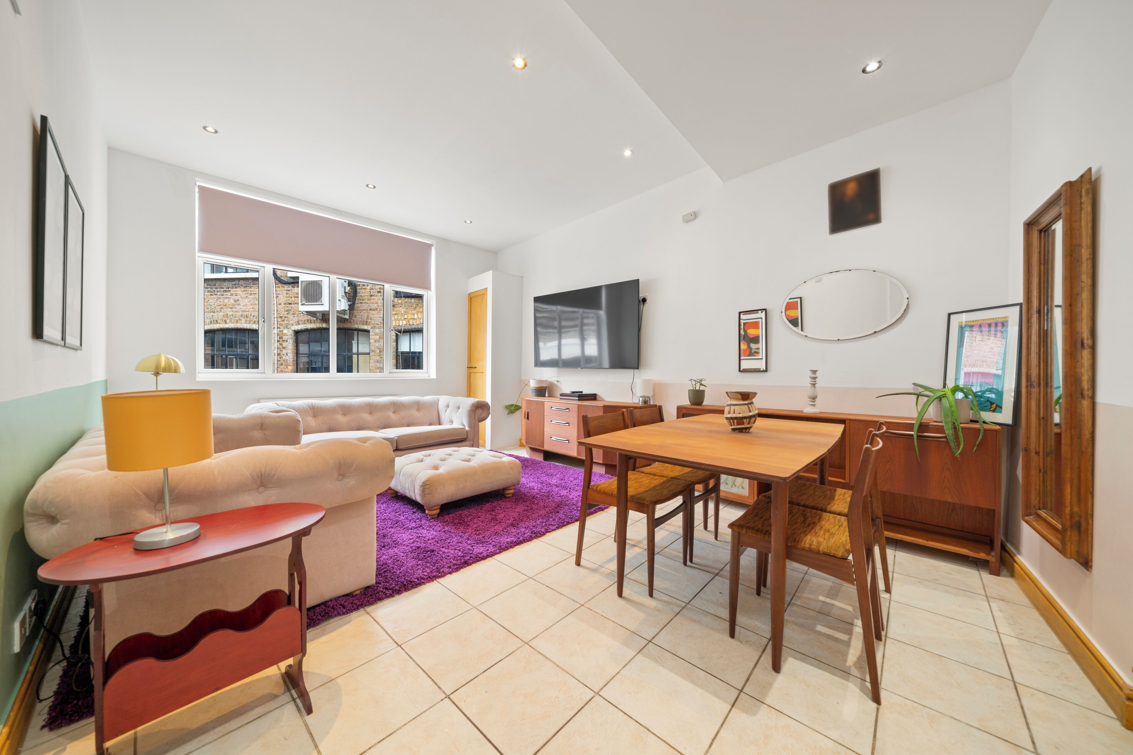 Uma sala de estar moderna com sofá, puf e mesa de jantar com cadeiras. Há uma TV na parede e uma janela que oferece luz natural.