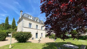 Un grand bâtiment historique à la façade symétrique, doté d’une entrée centrale et d’un jardin bien entretenu avec une pelouse et un espace aménagé pour s’asseoir.