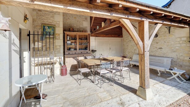 Un bâtiment en pierre agrémenté d’une pergola en bois, une table à manger avec des chaises, un banc et une petite table sur laquelle est posé un vase.