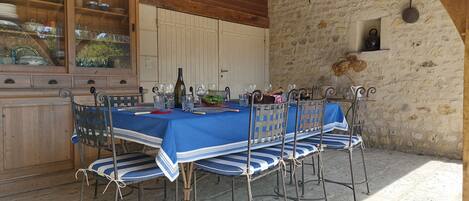 A rustic outdoor dining area with a stone wall, wooden ceiling beams, and metal chairs with blue and white striped cushions.