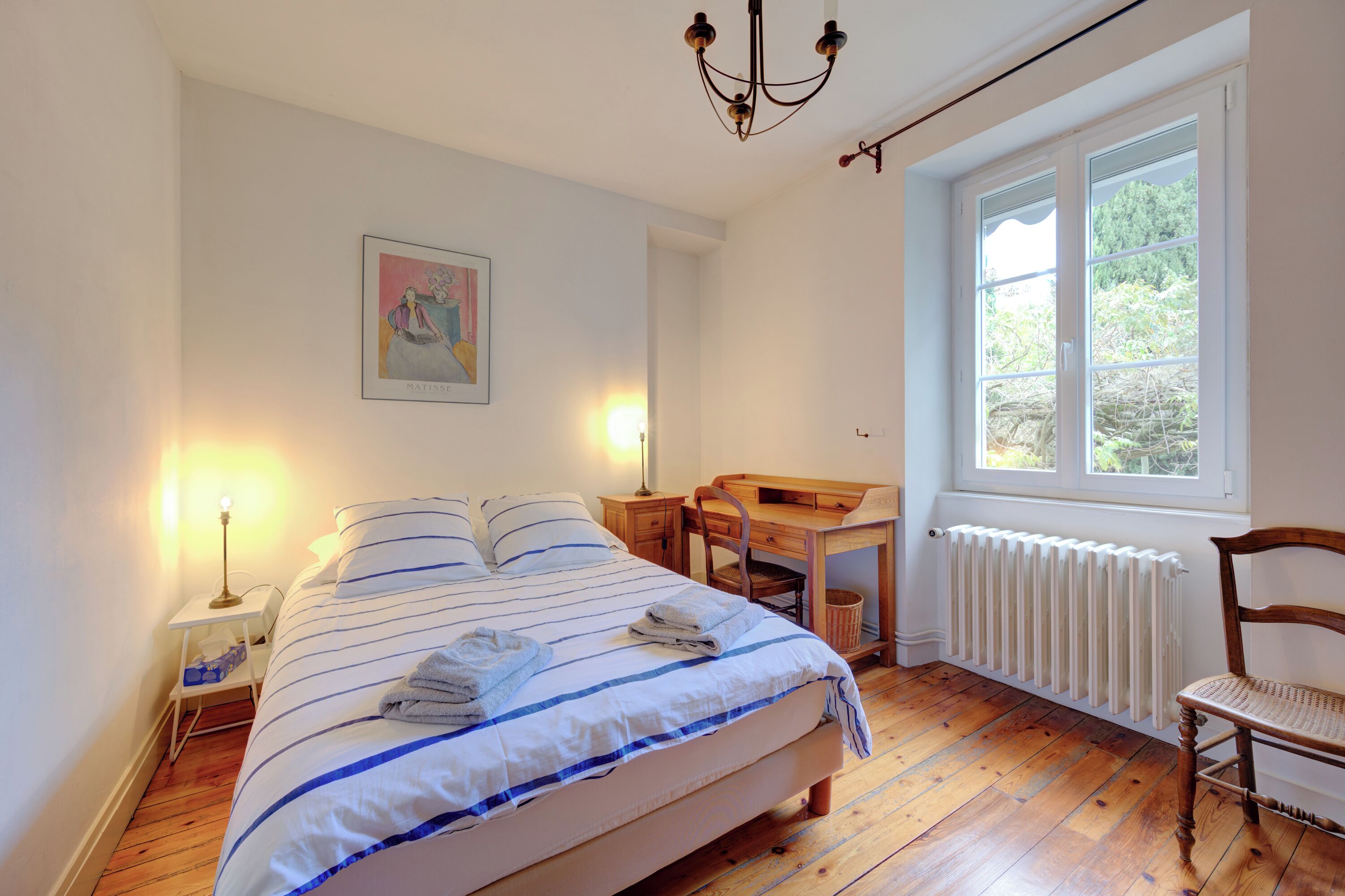 Un dormitorio con cama, mesita de noche, lámpara, escritorio, silla, radiador y ventana con vistas al verdor.