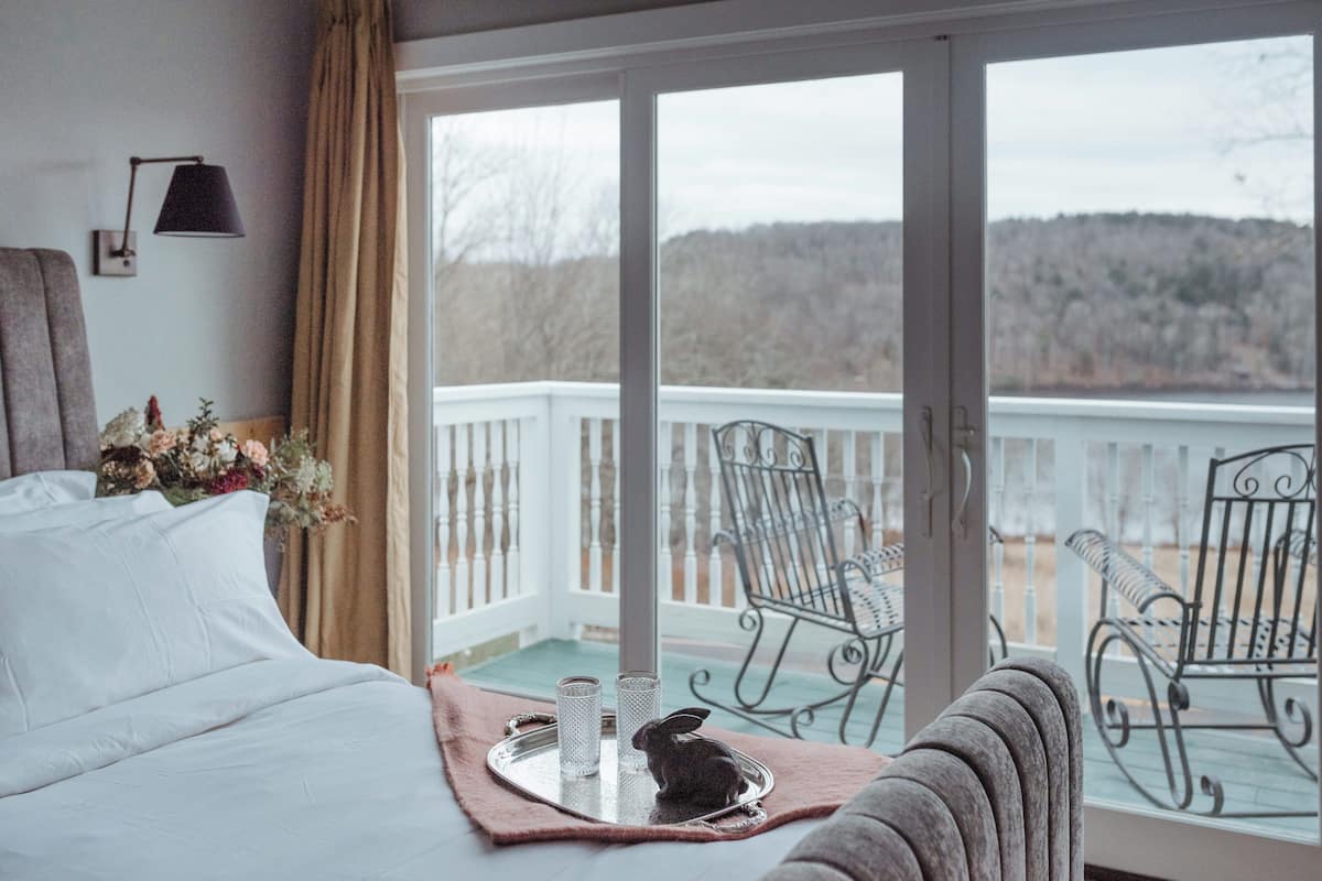 Un dormitorio con cama, una mesita de noche con lámpara, una ventana con vistas a un balcón, y una silla en el balcón.