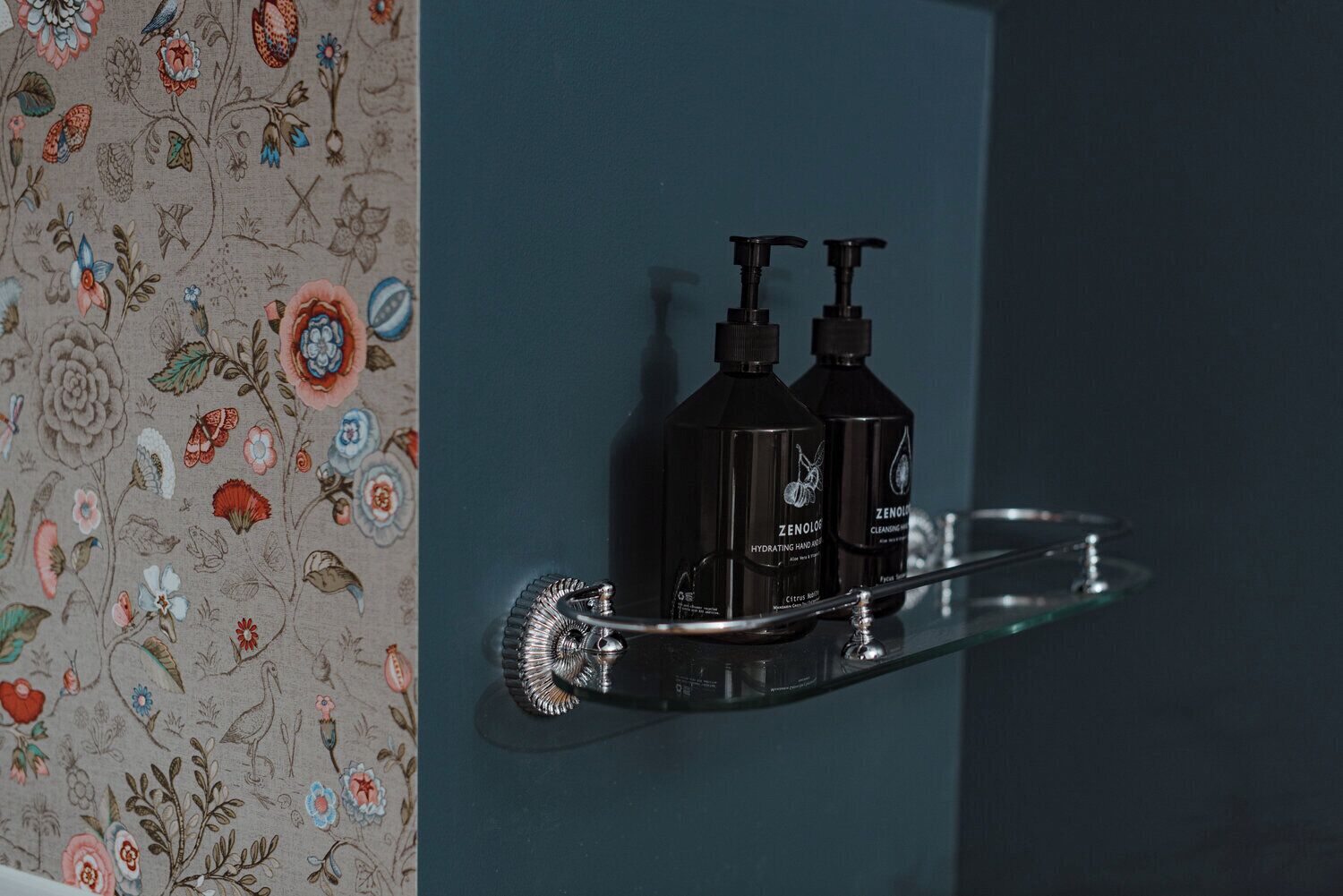 A bathroom with a floral wallpaper and a shelf holding two black bottles labeled 'ZENOLIS HYDRATING HAND CARE'.