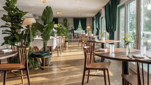 Un amplio comedor con varias mesas y sillas, grandes macetas con plantas y vistas al exterior a través de grandes ventanales.