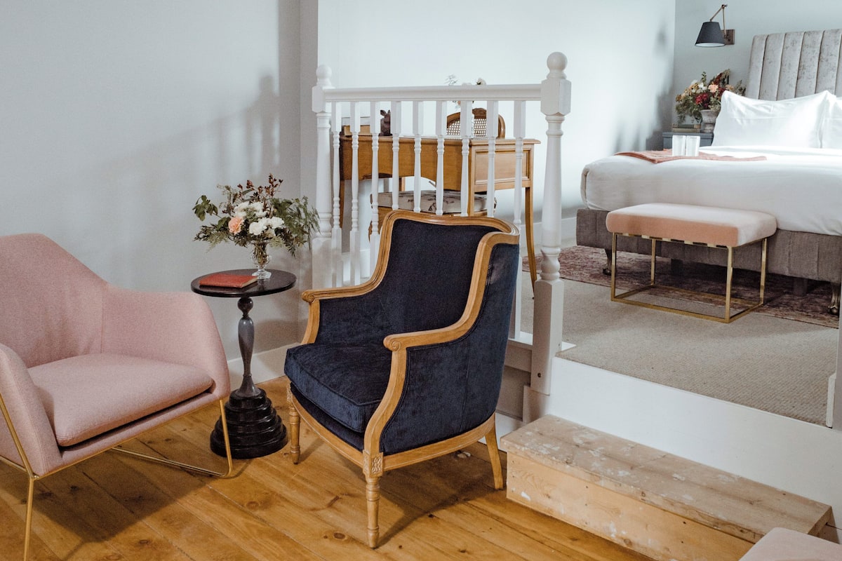 Una habitación con piso de madera, una cama con ropa blanca, un sillón tapizado en azul, un sillón rosa y una mesita redonda con un florero.