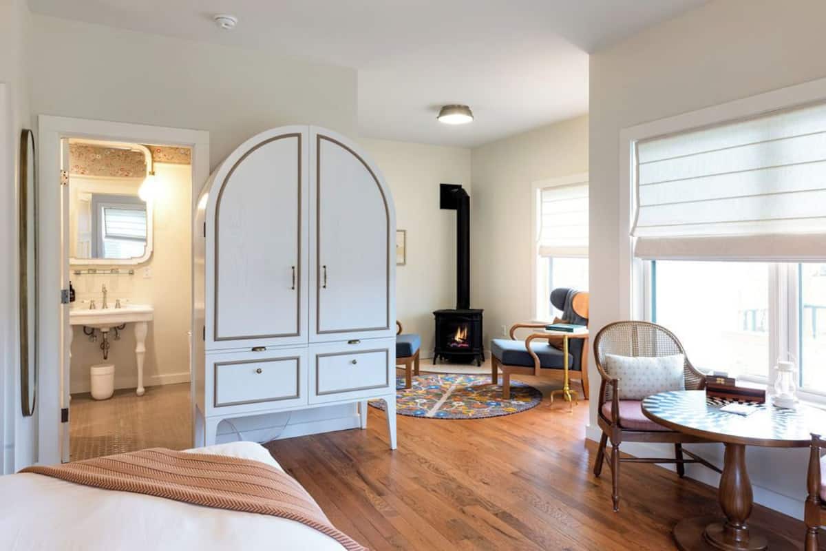 Un dormitorio con cama, armario, tocador, chimenea, una mesa redonda y ventana con persianas.