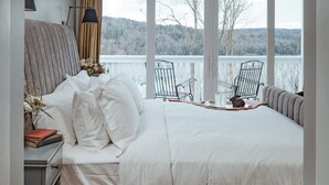 Un dormitorio con una cama grande, una mesita de noche con libros, una lámpara y vistas a un lago y árboles a través de amplias ventanas.