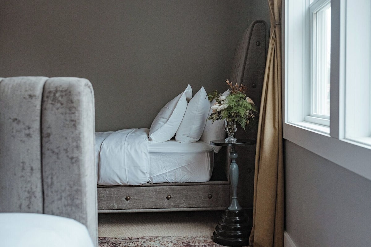 Un dormitorio con cama, una mesita de noche con un florero, una silla y un ventanal con cortinas.