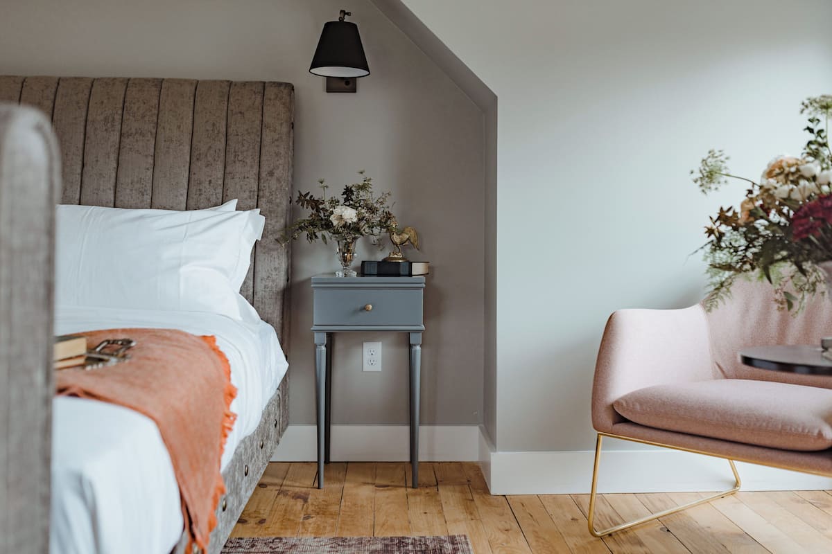 Un dormitorio con cabecera de madera, una mesita de noche con lámpara, una silla y un arreglo floral.