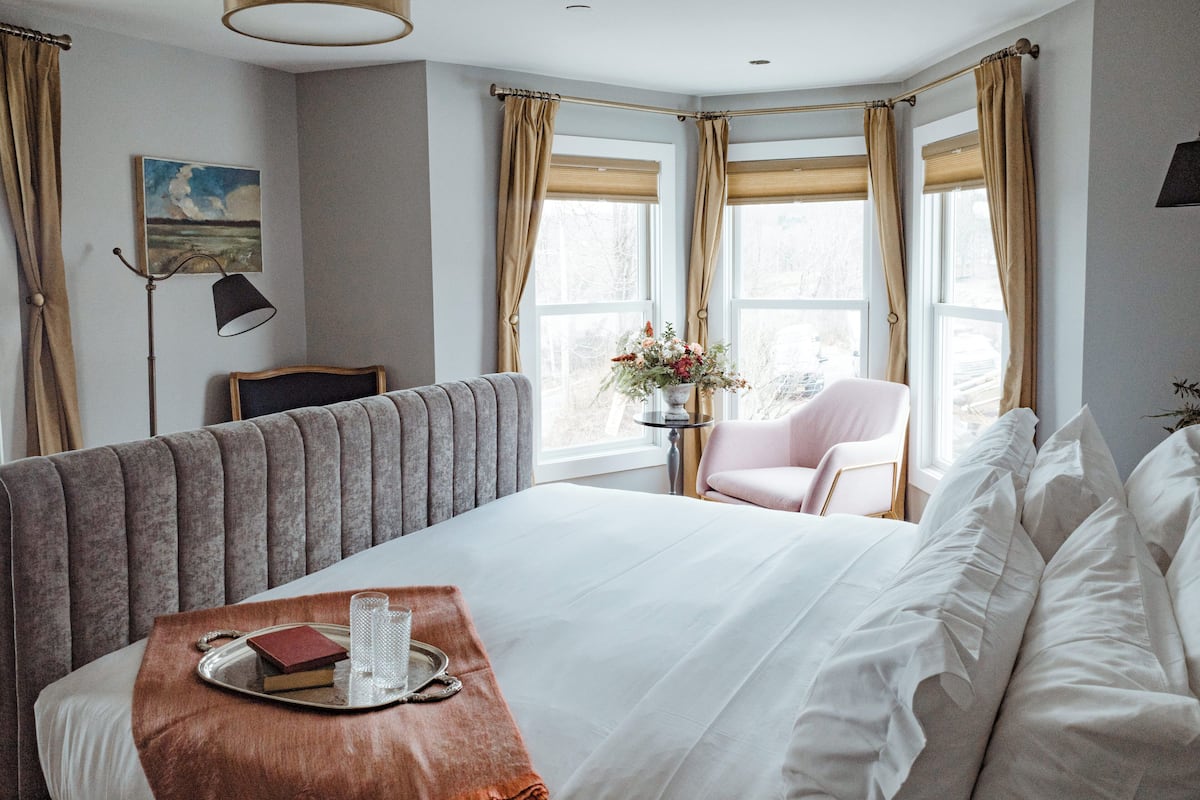 Habitación de hotel ordenada con una cama grande, una mesita de noche con bandeja, una silla y una pequeña mesa con flores.