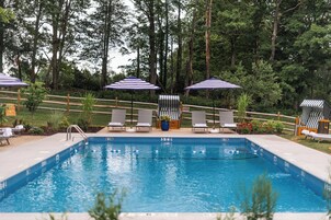 Zona de piscina con sillones de descanso y sombrillas a rayas, rodeada por una cerca de madera y árboles.