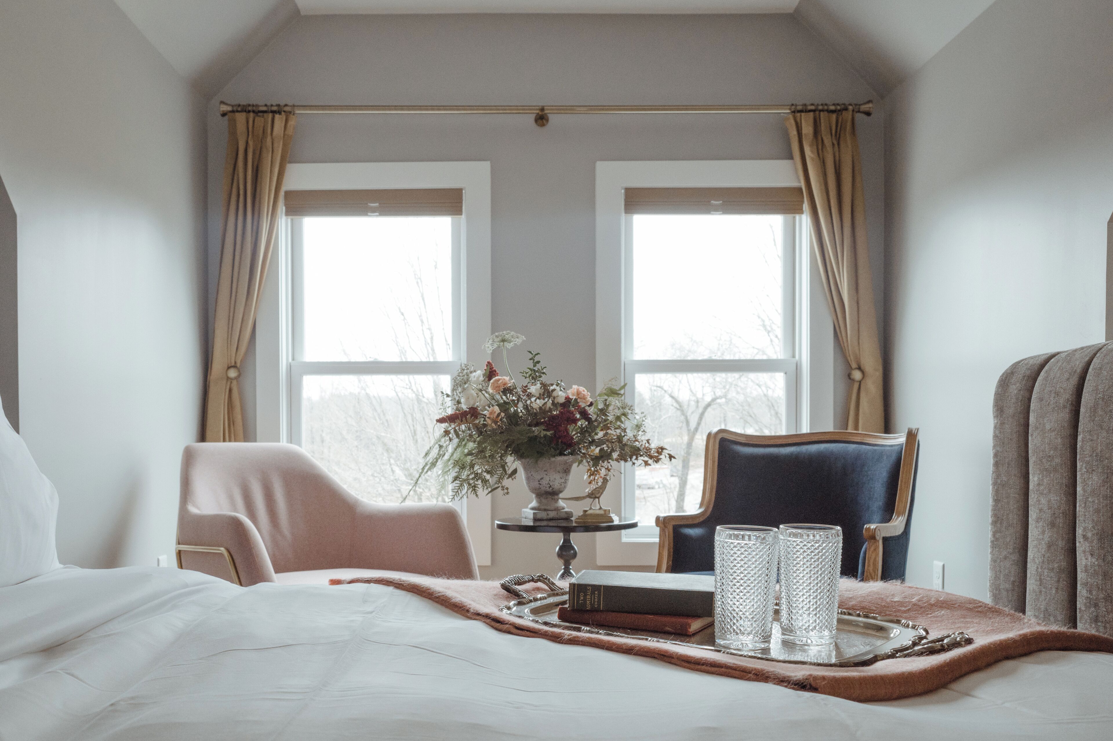 A bedroom with a bed, two chairs, a table with a vase of flowers, and two glasses.