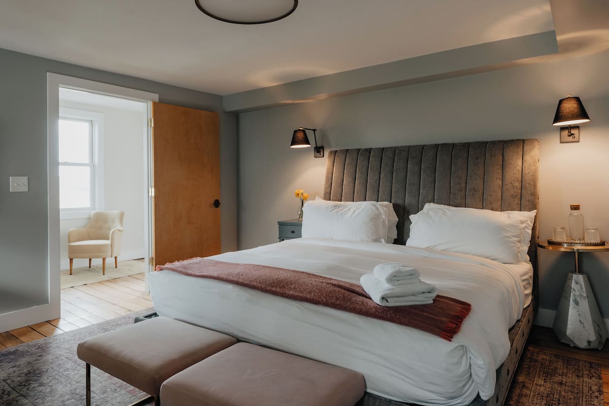 Un dormitorio moderno con una cama grande, lámparas de noche, una mesita de noche con un florero y una puerta que da a otra habitación.