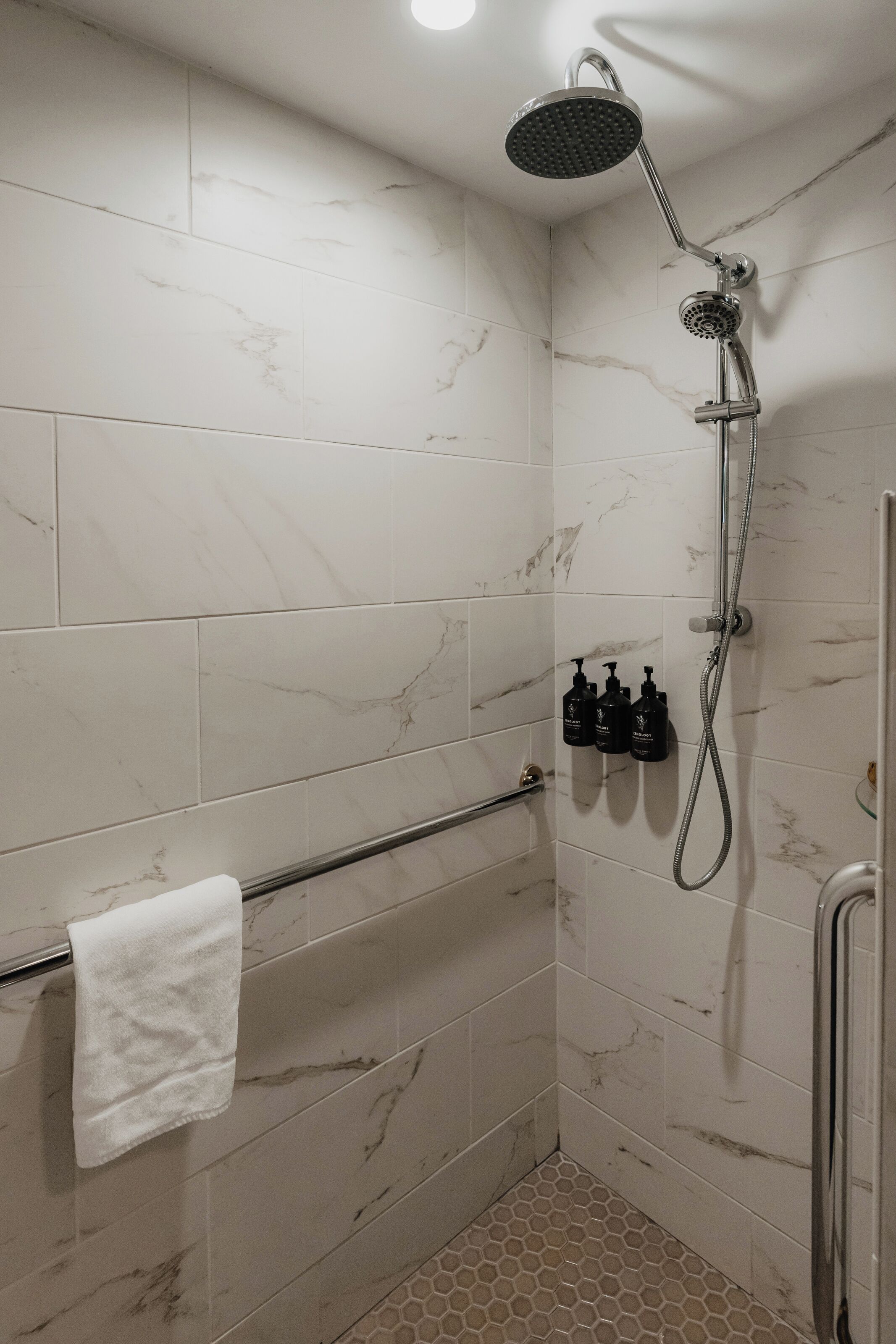 A shower area with a handheld showerhead, a towel rack with a white towel, and three bottles of shampoo.
