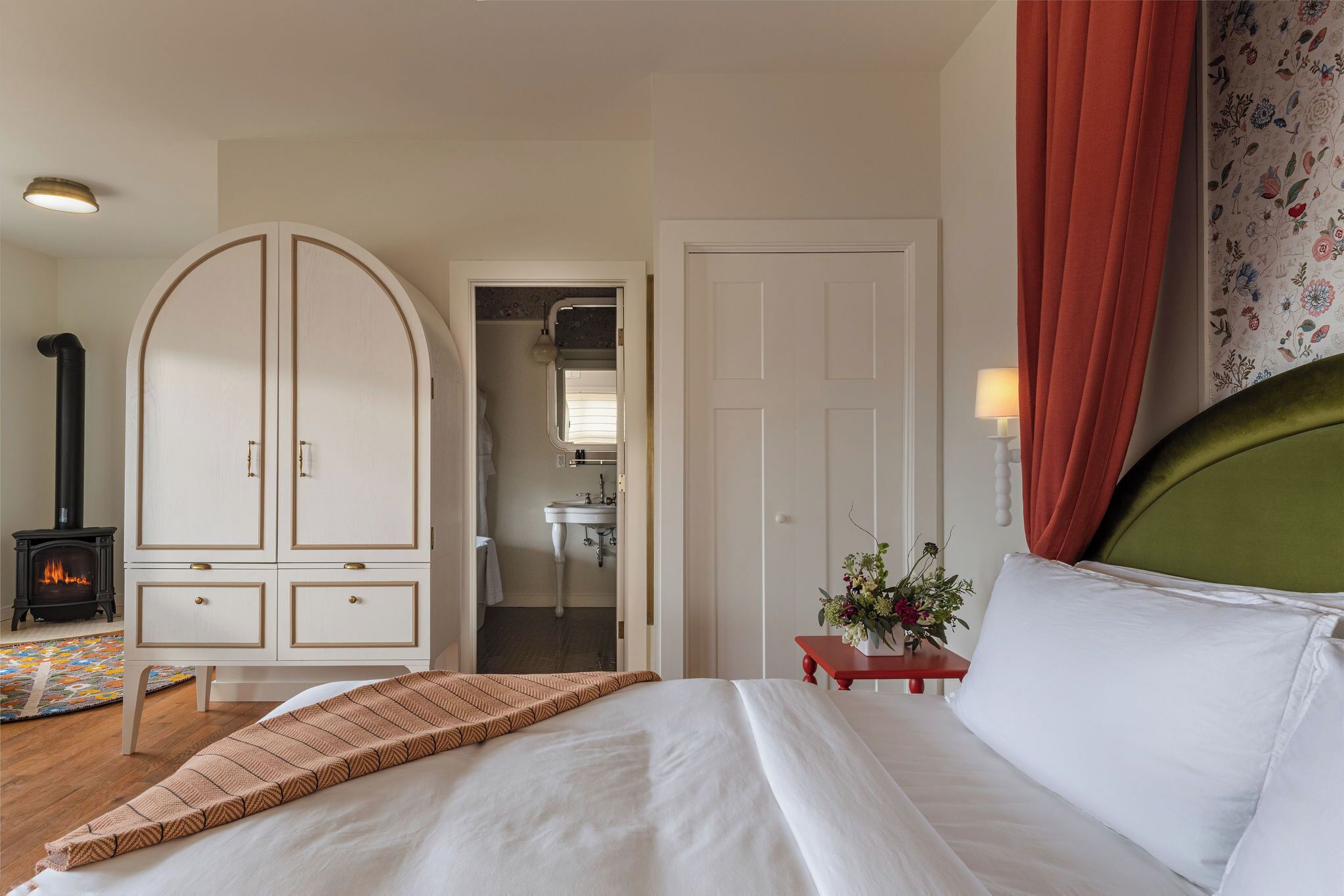 A bedroom with a bed, a wardrobe, a nightstand, a lamp, and a view of a bathroom.