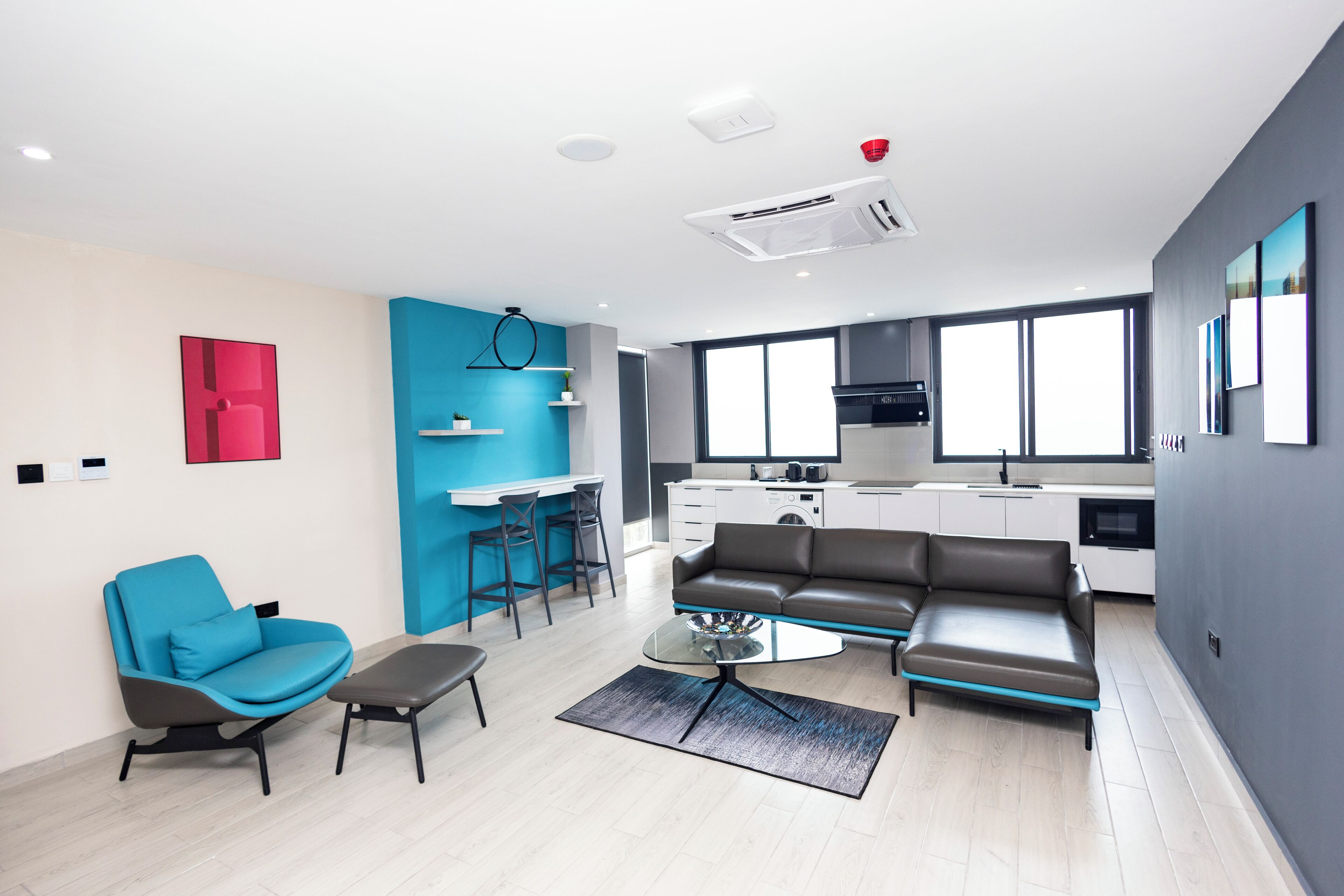 A modern living room with a sofa, a glass coffee table, and a blue armchair. There is a kitchen area with a stove and a sink.
