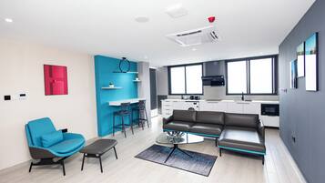 A modern living room with a sofa, a glass coffee table, and a blue armchair. There is a kitchen area with a stove and a sink.