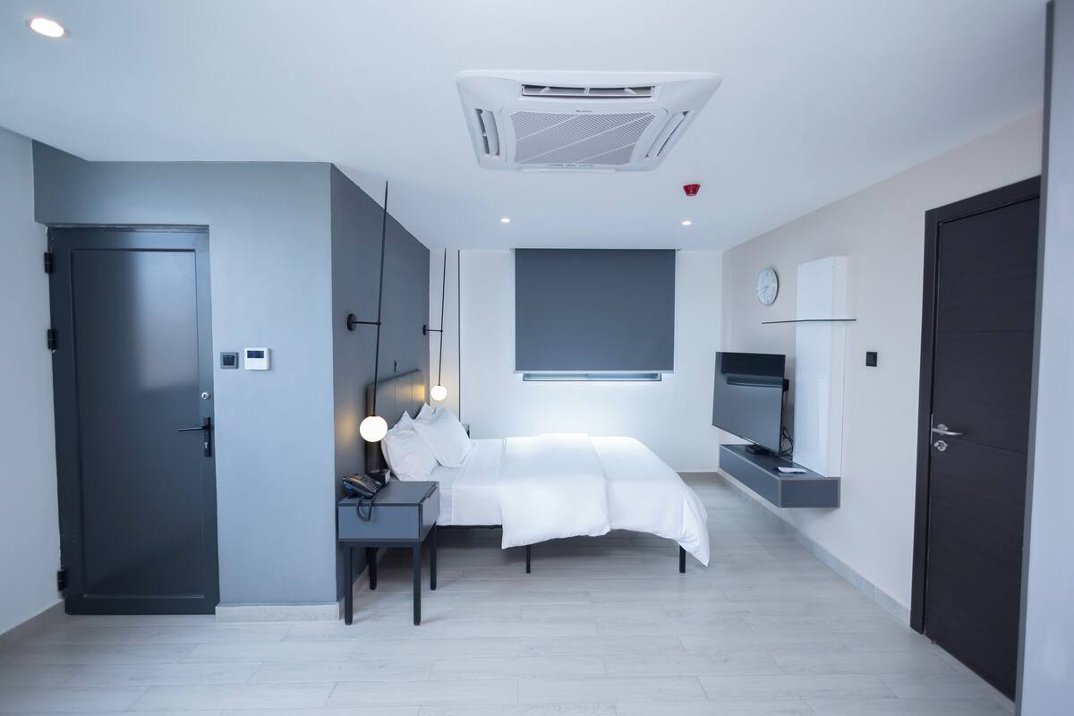 A modern hotel room with a bed, bedside table, lamp, air conditioning unit, and a TV mounted on the wall.