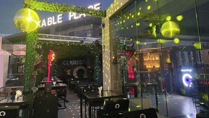 An outdoor dining area with tables and chairs, a stage with a 'CLOUD9' sign, and a glass wall reflecting lights. - Bankable Roomzzz (Accra)