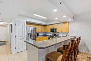 A modern kitchen with granite countertops, stainless steel appliances, wooden cabinets, and a bar area with bar stools. - 🌊OW14-204 | Pet and Family-Friendly Oceanwalk Condo Near Pool & Beach🐶 (New Smyrna Beach)