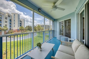 A balcony with a ceiling fan, a seating area with a sofa and a small table, and a view of a residential area with buildings and greenery.