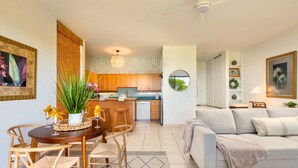 A modern kitchen with wooden cabinets, a dining area with a round table and chairs, and a living room with a sofa and a round mirror. - NEW! Big Island Stay with Private Lanai, Golf Course, Pool, Pickleball, & Views! (Waikoloa Village)