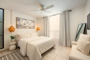 A bedroom with a bed, a ceiling fan, a large window with curtains, a side table with a plant, and a painting on the wall.