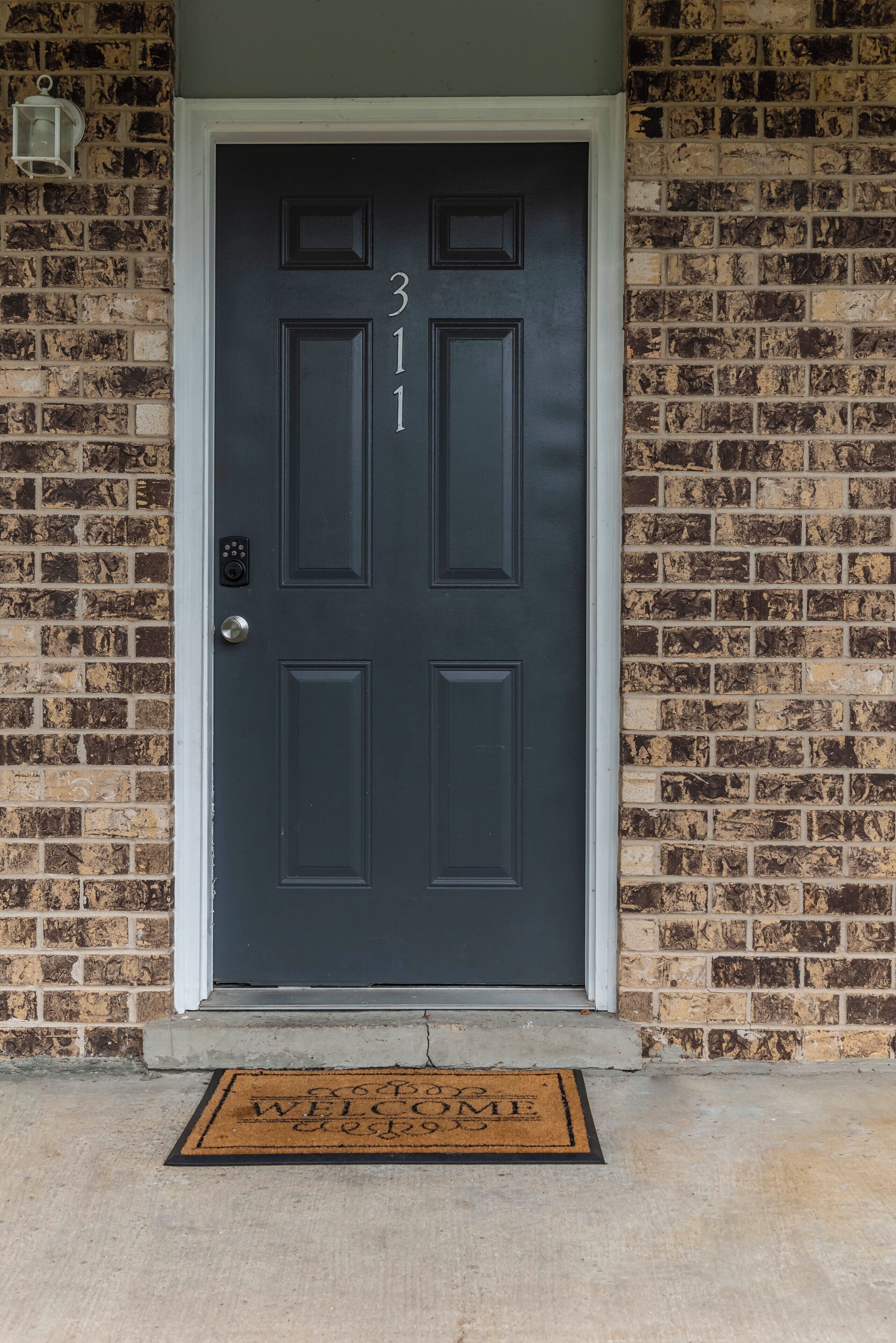 A dark-colored door with the number 311, a metal doorbell, and a doormat with "WELCOME HOME" text.