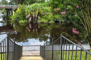 Een houten trap die leidt naar een brug over een kalm waterlichaam, omgeven door weelderige begroeiing en roze bloemen.