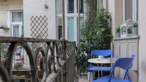 Un balcon avec une balustrade en métal, deux chaises bleues, une petite table et un pot de fleurs.