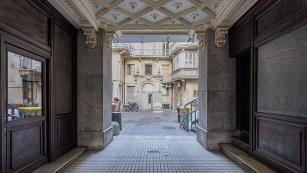 Un couloir orné de moulures au plafond et doté de grandes portes en bois, qui mène à une cour intérieure avec une fontaine et des vélos.
