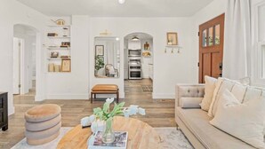 Amplia sala de estar con sofá, reposapiés y una mesa de centro, una cocina al fondo y un comedor con candelabro.